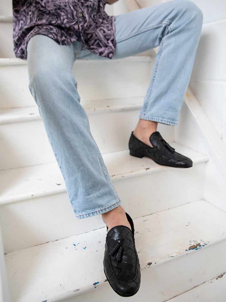 Aidan Black Woven Leather Loafers with Leather Tassels, styled with light-wash denim jeans, worn by a seated model on white stairs - POEVE Collection