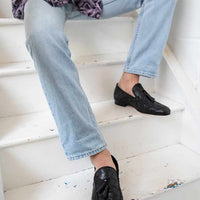 Aidan Black Woven Leather Loafers with Leather Tassels, styled with light-wash denim jeans, worn by a seated model on white stairs - POEVE Collection