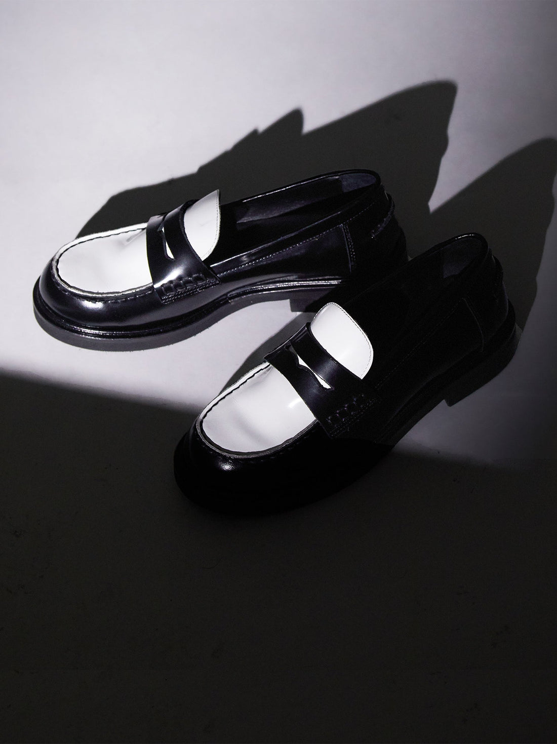 Ollie Black and White Brushed Leather Penny Loafers displayed under a dramatic spotlight, casting bold shadows on a minimalist grey surface - POEVE Collection