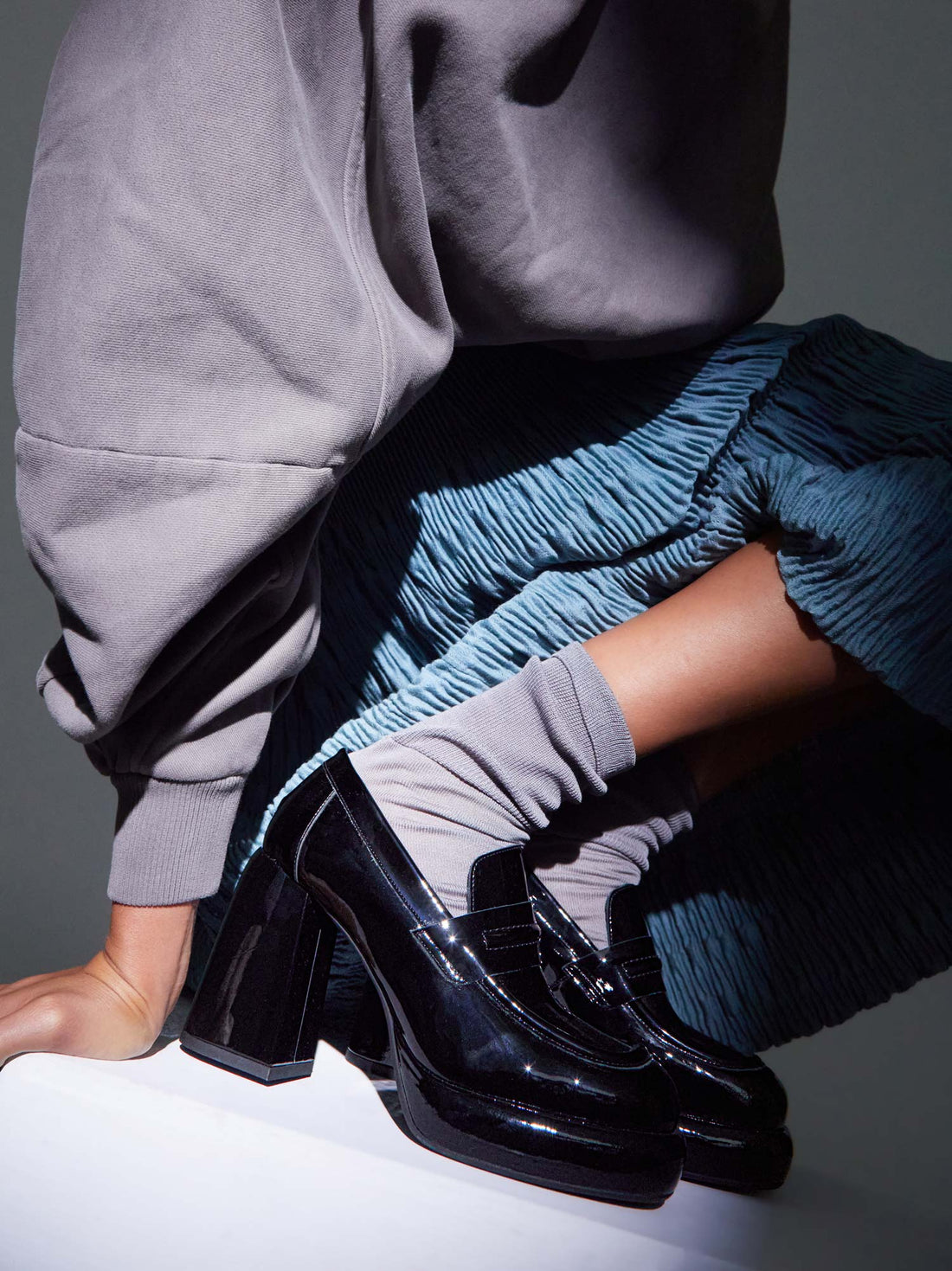 Zery Black Patent Leather Heel Loafers, styled with grey slouchy socks, a pleated blue skirt, and an oversized grey sweatshirt - POEVE Collection