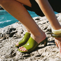 Mavis Green Crinkled Patent Leather Heel Mules worn barefoot, styled with a black dress, against a rocky seaside backdrop - POEVE Collection